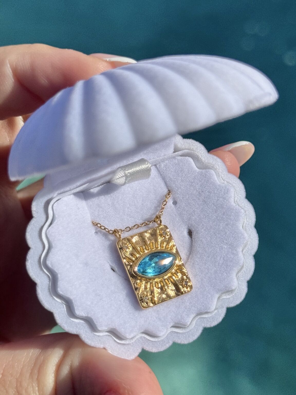 A gold pendant necklace with a marquise-cut light blue gemstone, displayed inside a white clamshell jewelry box held by a hand against a blurred teal background.