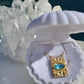 A gold pendant necklace with a rectangular gold frame and a light blue oval gemstone, displayed inside an open white clamshell jewelry box. The box is set against a background of clear quartz crystal points and a dark blue surface.
