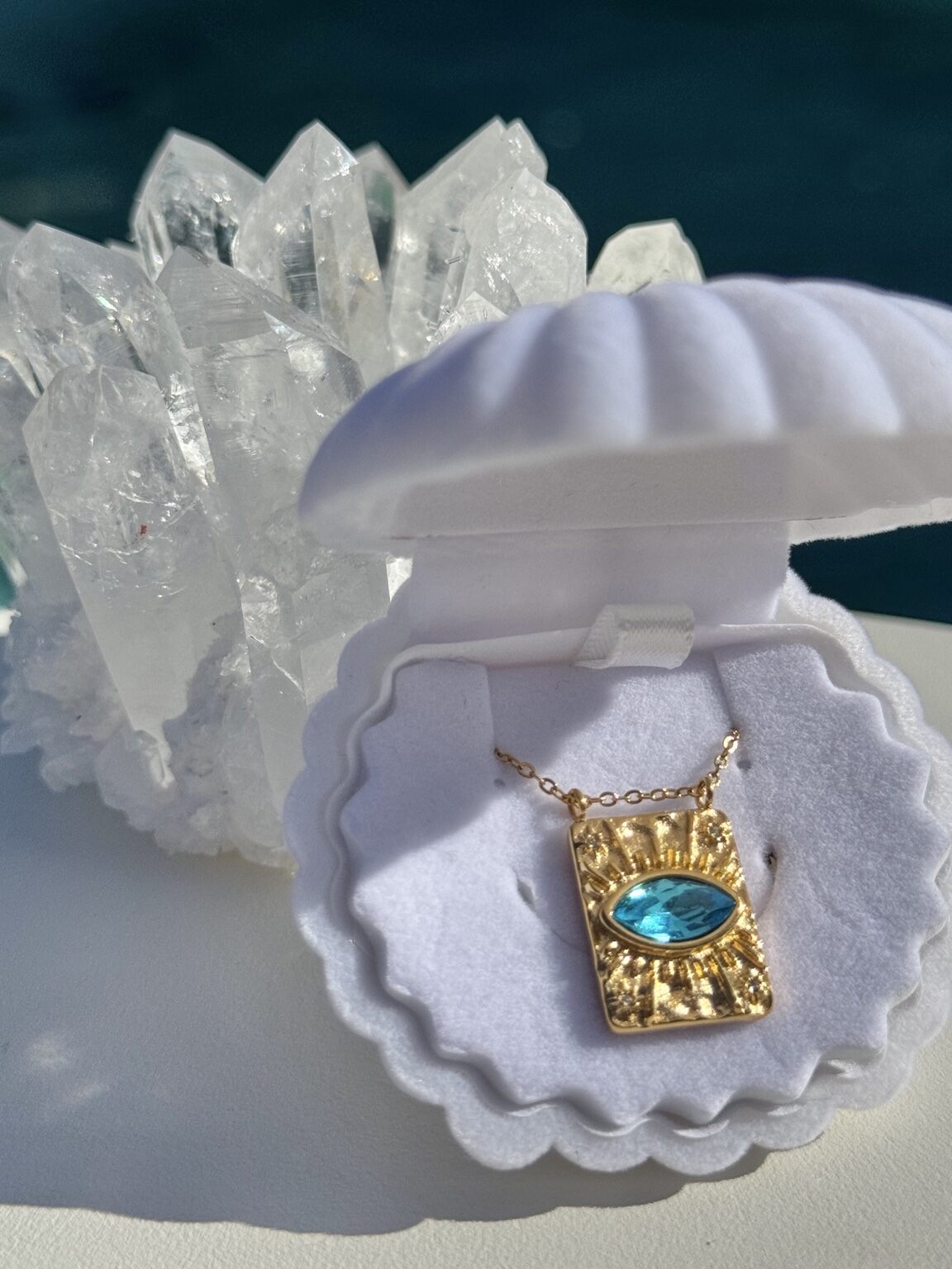 A gold pendant necklace with a rectangular gold frame and a light blue oval gemstone, displayed inside an open white clamshell jewelry box. The box is set against a background of clear quartz crystal points and a dark blue surface.