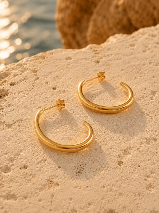 A pair of gold hoop earrings with small flower-shaped posts, resting on a textured beige stone surface with a blurred outdoor background.