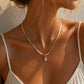 A close-up lifestyle shot of a model wearing a delicate gold snake chain necklace with a small clear rectangular crystal pendant, styled with a white spaghetti strap top under warm sunlight.