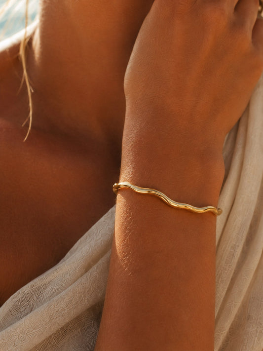 A close-up of a person's wrist wearing a thin, wavy gold bangle bracelet, with warm sunlight and a light fabric draped nearby.