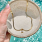 A gold snake chain necklace with a small square-cut clear gemstone pendant, held in a speckled beige ceramic bowl over turquoise water.