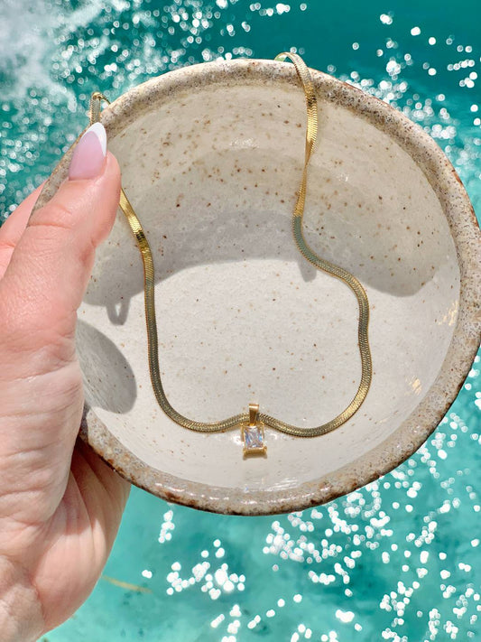 A gold snake chain necklace with a small square-cut clear gemstone pendant, held in a speckled beige ceramic bowl over turquoise water.