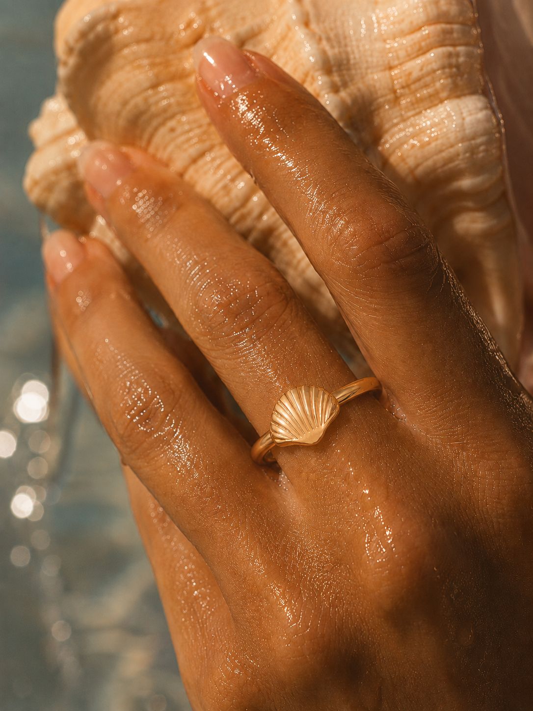 A close-up of a hand with wet skin wearing a delicate gold ring featuring a conch shell, holding a large seashell against a blurred water background.