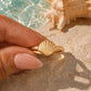 A gold ring with a seashell design held between fingers, set against a background of a pool and a large seashell on a sandy surface. Conch Shell