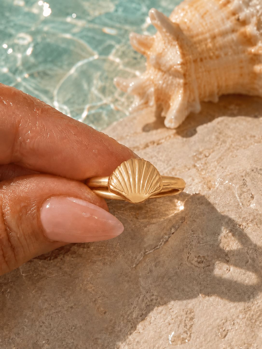 A gold ring with a seashell design held between fingers, set against a background of a pool and a large seashell on a sandy surface. Conch Shell