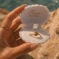 A hand holds an open clam-shell style ring box with the brand name 'Ocean The Label' printed inside. A gold ring shaped like a seashell is displayed in the center.