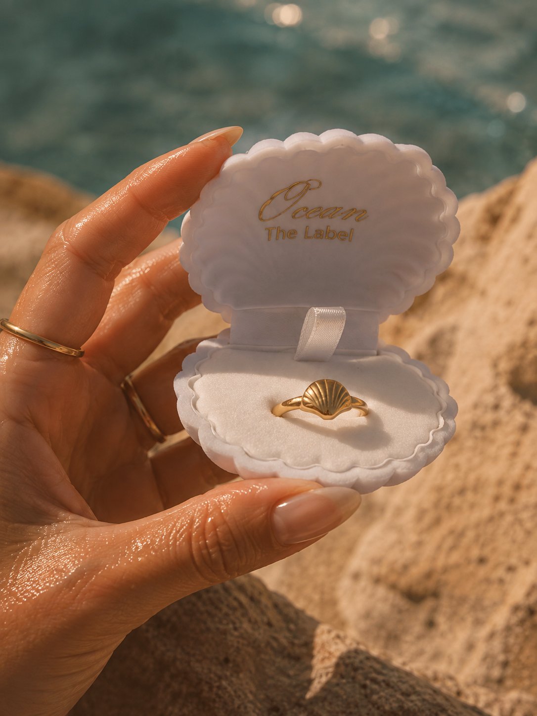 A hand holds an open clam-shell style ring box with the brand name 'Ocean The Label' printed inside. A gold ring shaped like a seashell is displayed in the center.