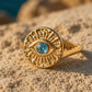 A gold signet ring with an evil eye motif and a central blue gemstone, resting on a sandy beach with a blurred ocean background.