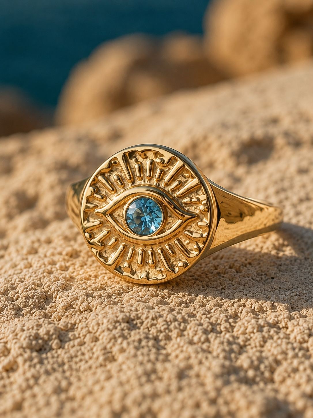 A gold signet ring with an evil eye motif and a central blue gemstone, resting on a sandy beach with a blurred ocean background.