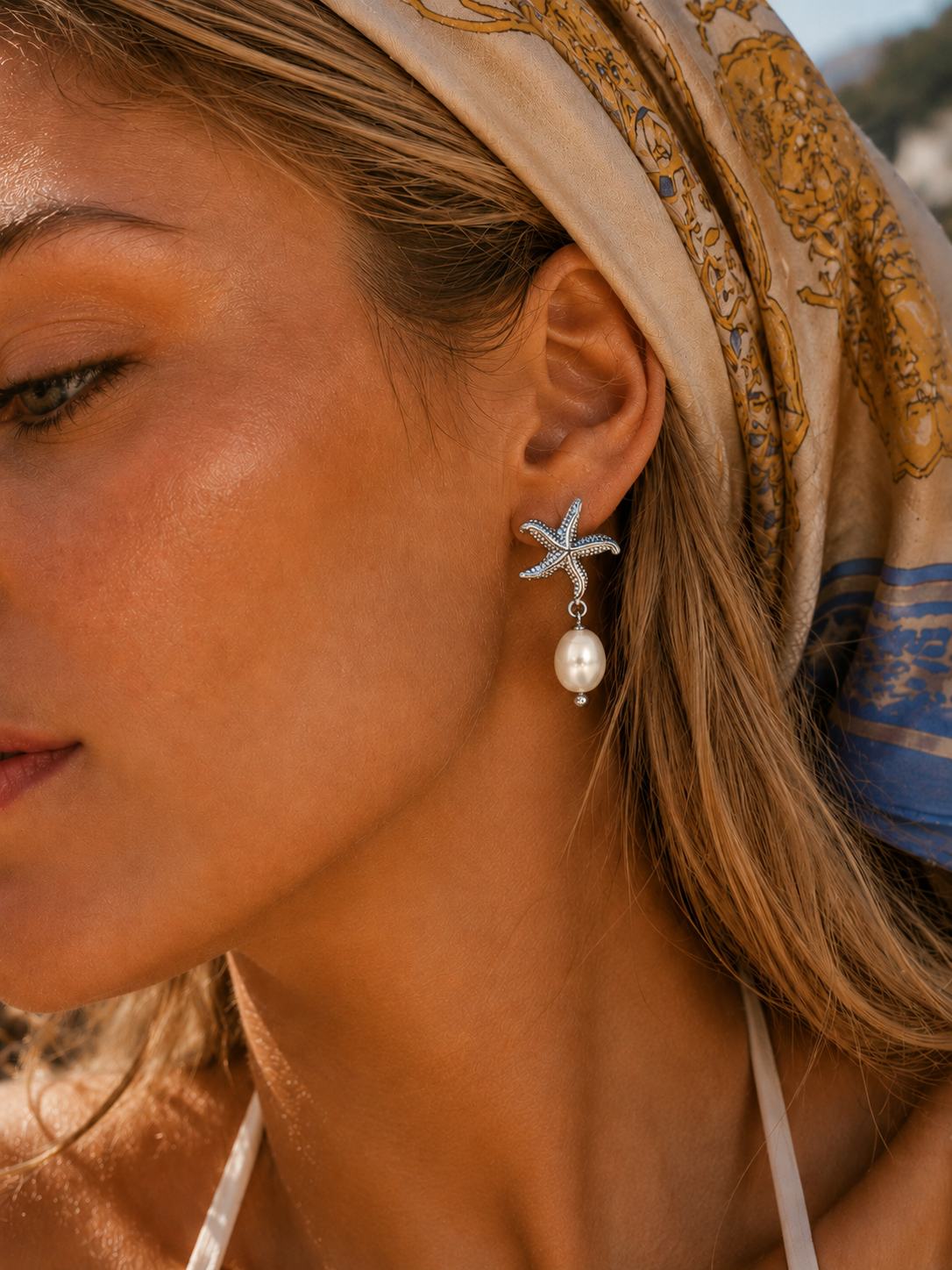 A close-up of a woman wearing a silver starfish dangle earring with a white pearl drop, styled with a patterned beige and blue headscarf.