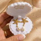 Gold starfish drop earrings with white pearl pendants, presented in a white clamshell jewelry box held by a hand on a sandy beach-themed background.