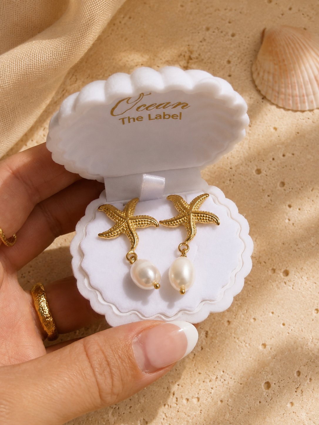 Gold starfish drop earrings with white pearl pendants, presented in a white clamshell jewelry box held by a hand on a sandy beach-themed background.
