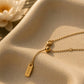 A delicate gold chain necklace with a small rectangular engraved tag reading 'OCEAN' and a small bead, displayed on a textured beige surface with warm sunlight and a blurred white flower in the background.