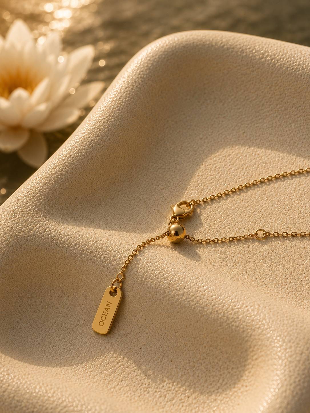 A delicate gold chain necklace with a small rectangular engraved tag reading 'OCEAN' and a small bead, displayed on a textured beige surface with warm sunlight and a blurred white flower in the background.