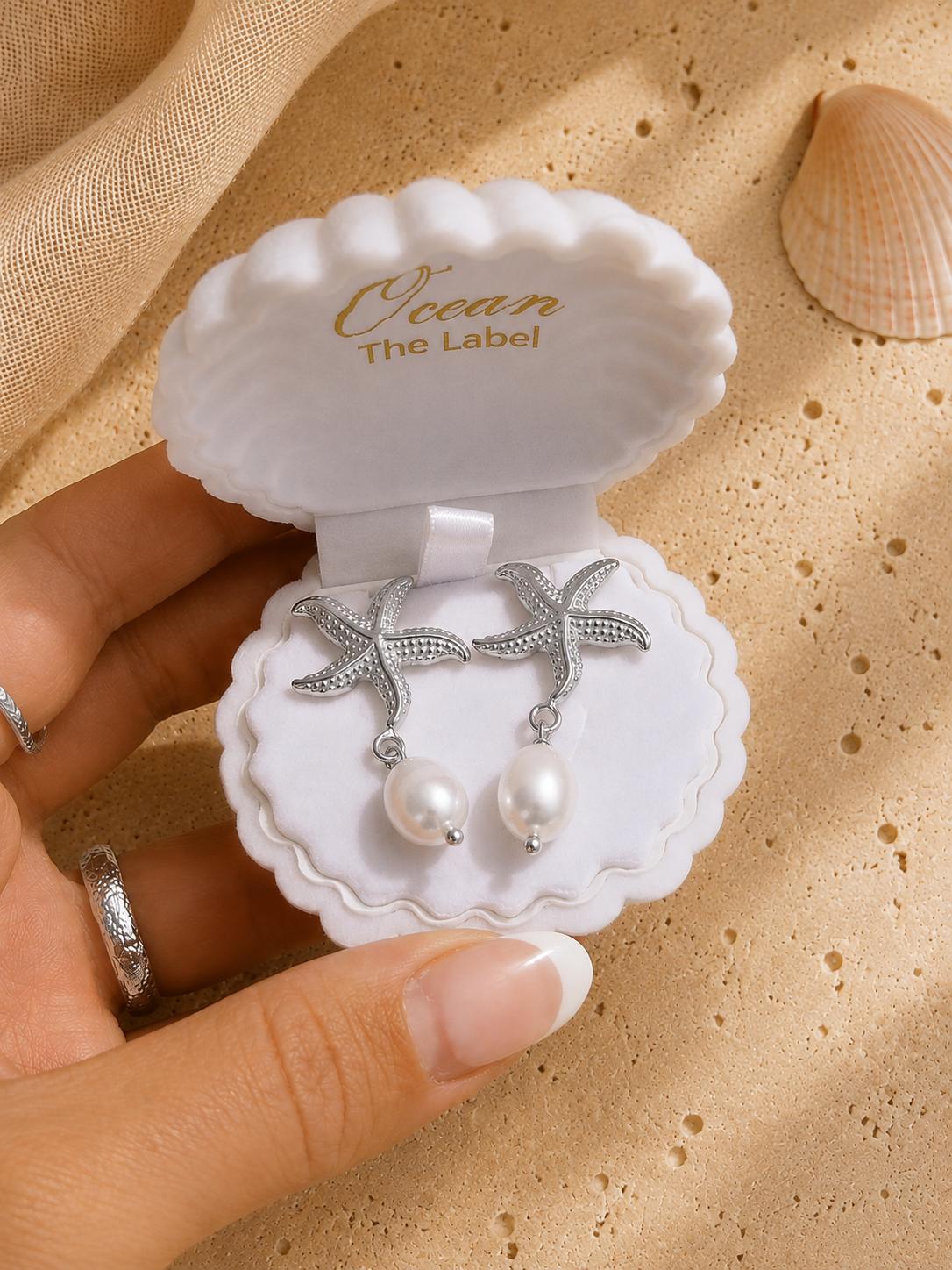 Maris silver starfish earrings displayed in a luxury flat lay, showing the detailed starfish design and pearl drops in clamshell box