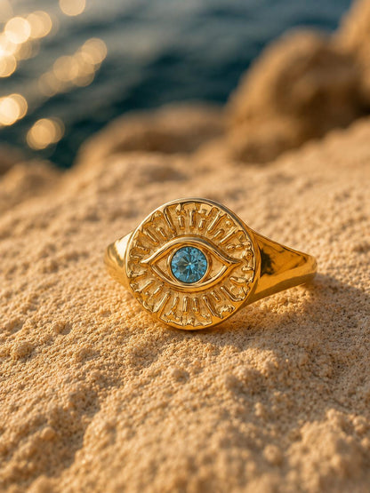 A gold signet ring with an eye motif and a blue stone in the center, resting on sandy beach ground with water in the background.