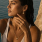 A woman in a white halter top wearing gold jewelry, including a delicate beaded necklace with a pearl-like pendant, a matching ring, and a small drop earring, posed outdoors near water and a rock with warm golden-hour lighting.