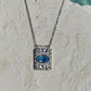 A silver protection energy rectangular pendant necklace with a textured sunburst evil eye design and a central oval blue zircon gemstone, hanging on a silver chain against a textured off-white stone background.