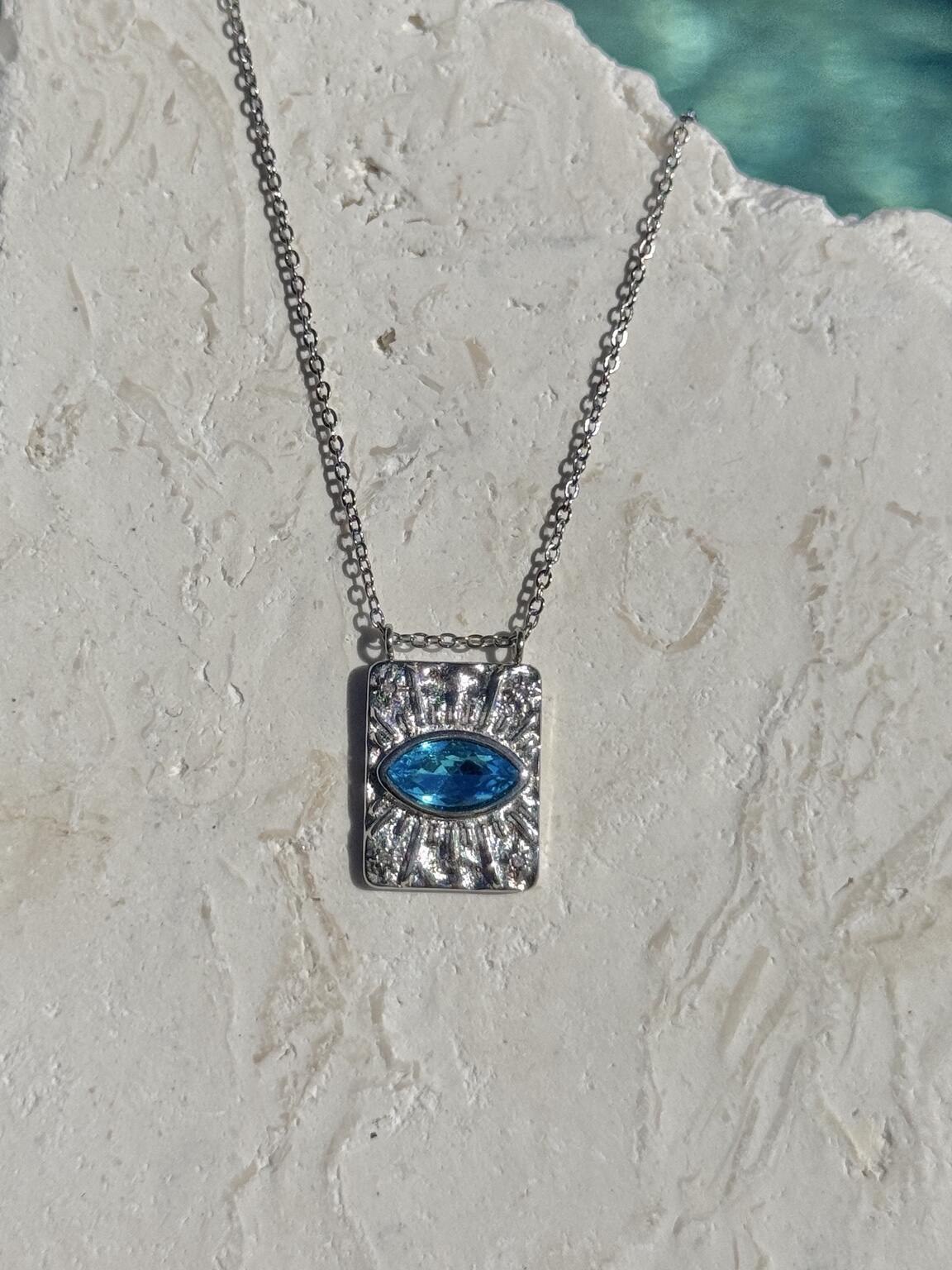 A silver protection energy rectangular pendant necklace with a textured sunburst evil eye design and a central oval blue zircon gemstone, hanging on a silver chain against a textured off-white stone background.