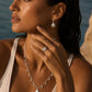 A woman wearing a silver sea shell necklace and matching drop earrings with white pearl-like beads, photographed outdoors near water with warm sunlight on her skin.
