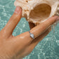 A silver seashell ring worn on a woman's finger, holding a large conch shell in clear turquoise water.