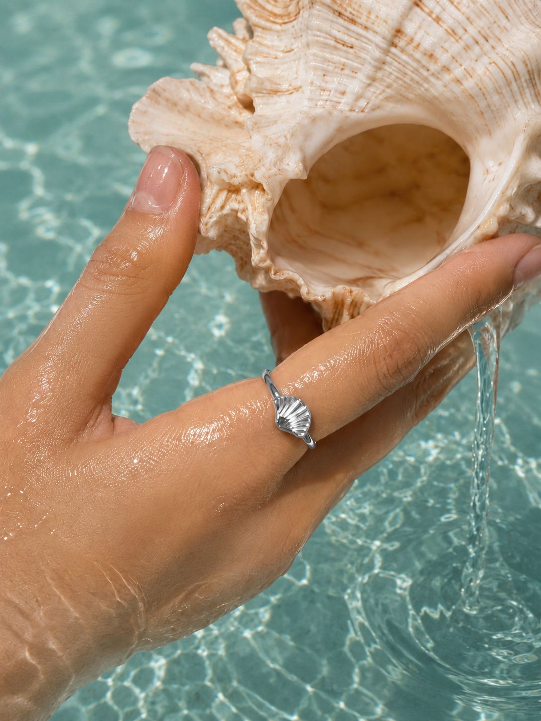 A silver seashell ring worn on a woman's finger, holding a large conch shell in clear turquoise water.